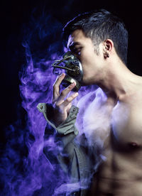 Shirtless man holding mask by smoke against black background