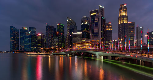 Illuminated modern buildings by river in city against sky at night