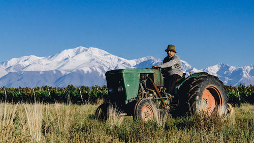 Man driving tractor on grassy field by vineyard against snowcapped mountains
