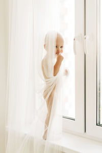 A little boy is standing on the windowsill near the window, hiding behind the curtain