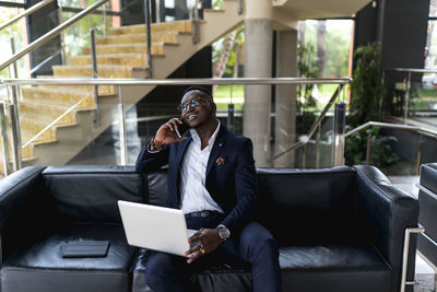 Handsome male businessman with laptop talking on phone call while sitting on sofa in hotel