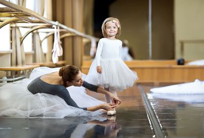 An adult ballerina is tying pointe shoes to a little ballerina.