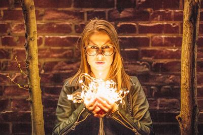 Portrait of young woman wearing eyeglasses holding illuminated string lights against brick wall