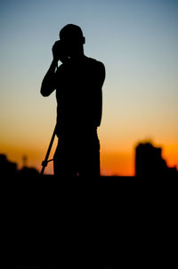 Silhouette man holding camera and standing against sky during sunset