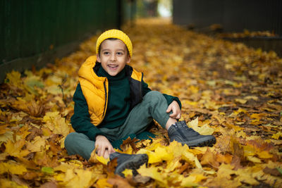 Portrait of a fashionable child boy autumn sitting on a trail in orange leaves in the afternoon