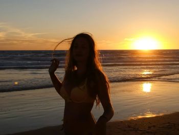 Portrait of young woman standing on beach against sky during sunset