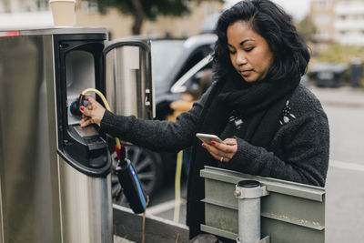 Mid adult woman using smart phone while charging car at station