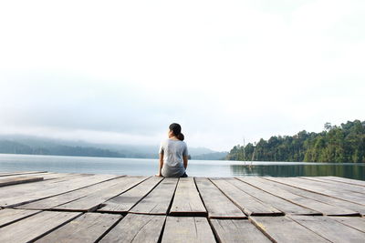 Rear view of man sitting on pier over lake against sky