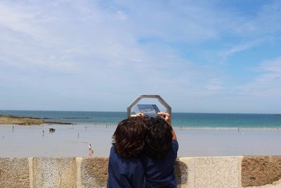 Rear view of brothers looking at sea through coin-operated binoculars against cloudy sky