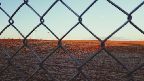 Windmills on landscape against clear sky seen through chainlink fence