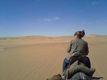 Rear view of woman sitting on camel at desert against blue sky