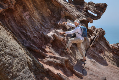 Man with camera and backpack climbing on rock formation against sky