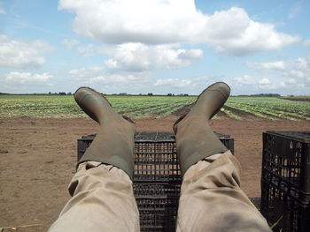 Low section of farmer legs on crate on agricultural field against sky