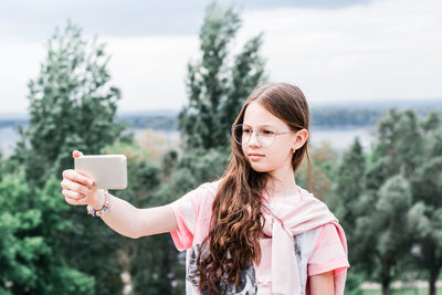 A serious girl in glasses takes a video selfie on a smartphone on the background of a river 
