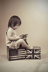 Side view of girl with rotary phone sitting on boxes with national flags over beige background