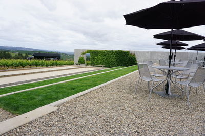 Chairs and tables under parasols by field against cloudy sky