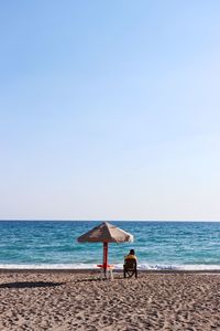 Rear view of man sitting on chair by parasol at beach against clear sky