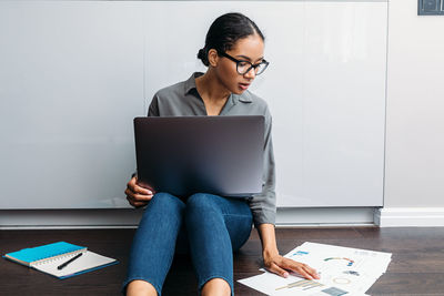 Young woman using laptop while sitting on floor against wall