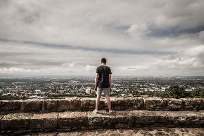 Rear view of man standing on retaining wall against cityscape