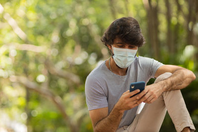 Young man wearing a mask and a smartphone at day time at a green park . mobile phone, technology