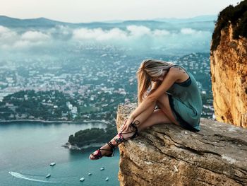 Side view of young woman sitting on mountain by sea against sky