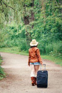 Rear view of woman pulling suitcase on dirt road against trees