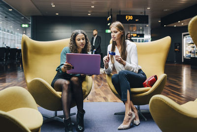 Full length of female multi-ethnic colleagues discussing over laptop and credit card while sitting at airport lobby