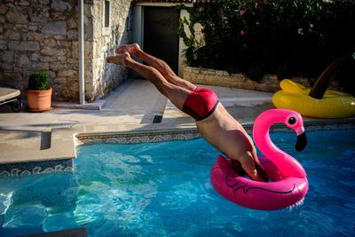 Shirtless mature man jumping on flamingo shape inflatable ring on pool