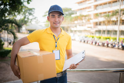 Portrait of smiling delivery man with package and clipboard standing at footpath 