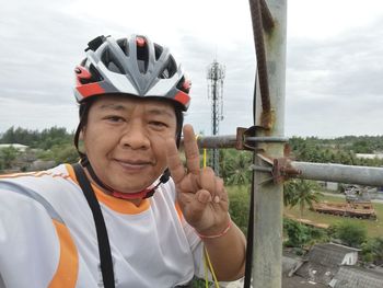 Portrait of man wearing cycling helmet gesturing peace sign against sky