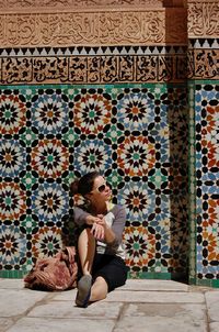 Woman in sunglasses sitting against patterned wall during sunny day