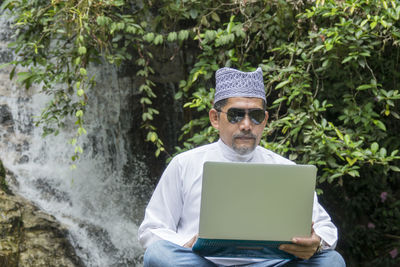 Mature man wearing sunglasses while using laptop against waterfall