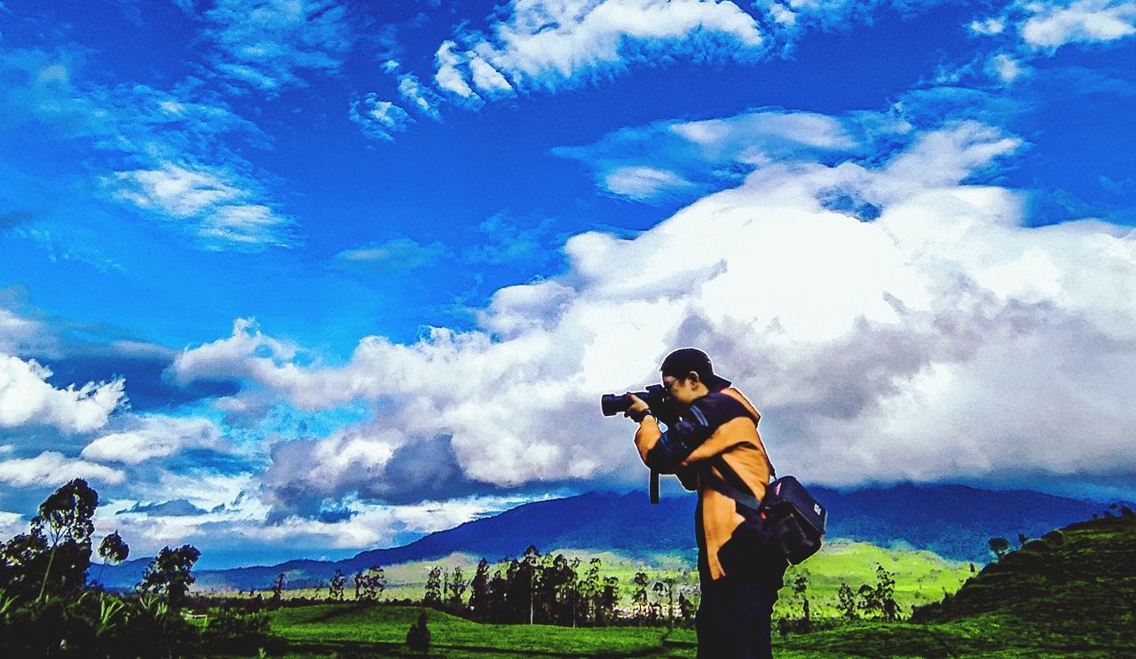 cloud, sky, mountain, one person, activity, nature, adult, leisure activity, men, landscape, beauty in nature, mountain range, sports, tree, standing, scenics - nature, environment, camera, outdoors, technology, occupation, blue, day, full length, land, lifestyles, photographing, person