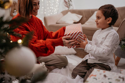 Mom giving gifts in boxes for the new year to her seven-year-old son near the christmas tree at home