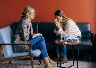 Crying depressed female client sitting on couch in therapist's workshop talking about trauma.