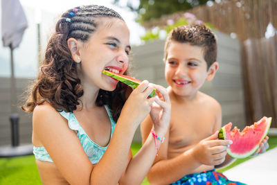 Two funny kids eating watermelon at the the edge of the pool on a summer day