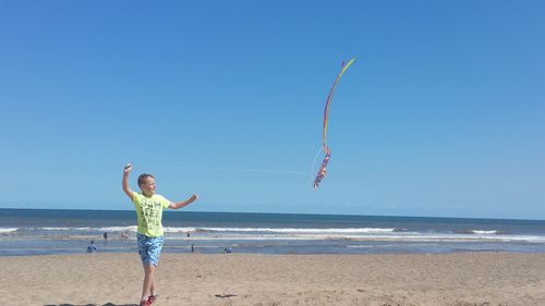 Full length of boy flying kite at beach against clear blue sky