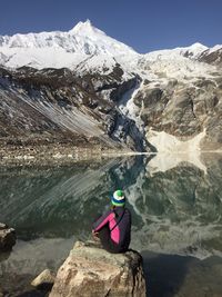 Rear view of woman sitting on rock by lake with snowcapped mountains reflection