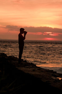 Silhouette man photographing sea against sky during sunset