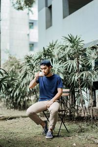 Full length of young drinking coffee while sitting on chair against plants