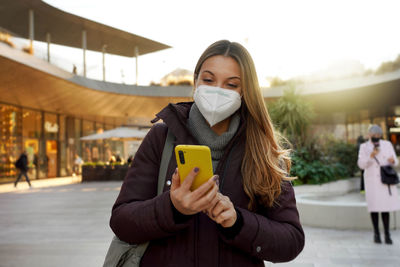Portrait of beautiful woman wearing medical face mask using internet wifi on phone in city at sunset