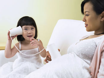 Girl listening pregnant mother abdomen with tin can phone on sofa