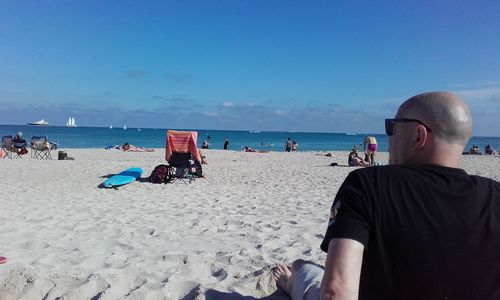 Rear view of man sitting at beach against blue sky