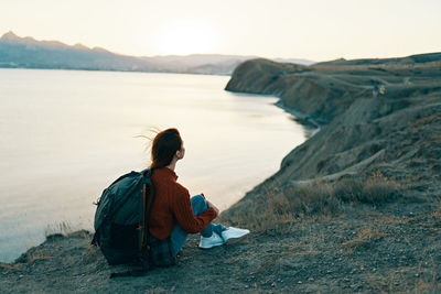 Man sitting on rock at beach against sky during sunset