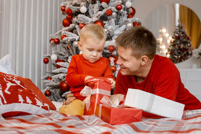 Father and son sitting on bed near christmas tree who is very busy unwrapping a present box