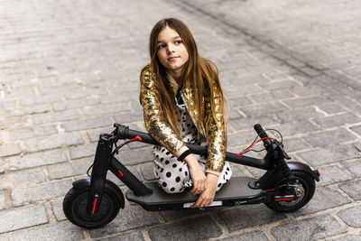 Portrait of girl wearing golden sequin jacket kneeling on folded e-scooter