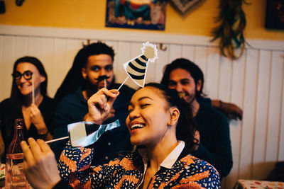 Cheerful young woman holding props while sitting against multi-ethnic friends at restaurant during dinner party
