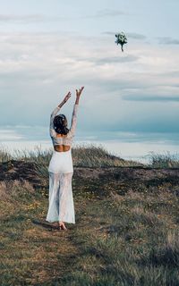 Rear view of bride throwing flower bouquet while standing on grass against sky