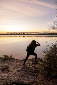 Rear view of woman standing on rock by lake against sky during sunset