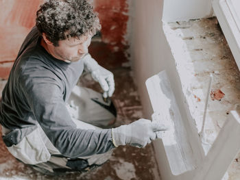 A young caucasian guy applies fresh putty with a large spatula on the wall under the window